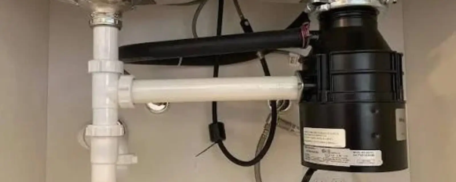 A garbage disposal mounted under a kitchen sink with drain pipes and hoses